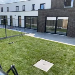 Świeżo skoszony trawnik przed nowoczesnym domem z czarnymi ramami okiennymi, widoczna czarna siatka ogrodzeniowa i szary właz kanalizacyjny na trawniku.