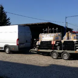 Biały bus z przyczepą przewożącą palety z czarnymi elementami, prawdopodobnie stalowymi, na tle budynku gospodarczego i domu jednorodzinnego.