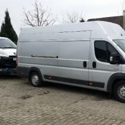 Srebrny bus ciągnie lawetę z białym dostawczakiem na tle brukowanej nawierzchni i zielonych krzewów.