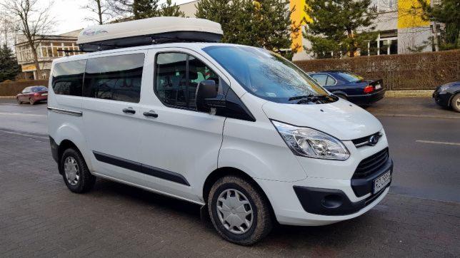 Biały van Ford Transit Custom z bagażnikiem dachowym na tle miejskiego krajobrazu z drzewami i budynkami.