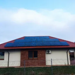 Dom jednorodzinny z panelami fotowoltaicznymi na czerwonym dachu, widoczne ogrodzenie z siatki i klimatyzator na ścianie budynku.