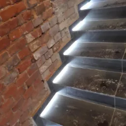 Schody z podświetleniem LED wzdłuż krawędzi stopni, przylegające do ściany z czerwonej cegły.