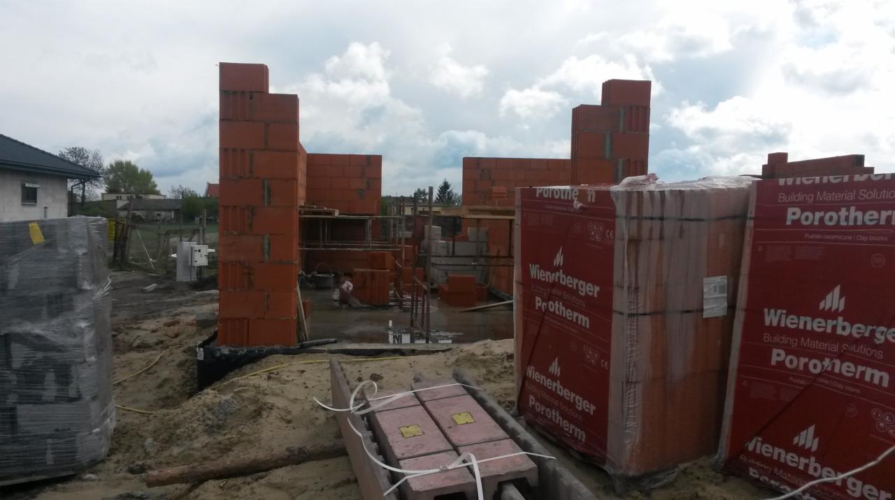 Wznoszone mury domu z czerwonej cegły Porotherm, widoczne palety z cegłami w folii, w tle budynki mieszkalne i zachmurzone niebo.