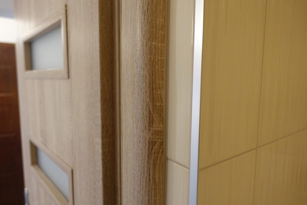 Fragment drzwi wewnętrznych z okienkami i ościeżnicą w kolorze jasnego drewna, widoczne płytki ceramiczne w tle.