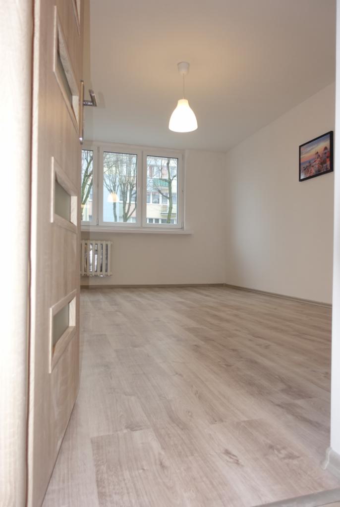 Jasne, puste pomieszczenie z panelami podłogowymi, widok zza uchylonych drzwi z przeszkleniami, okno z widokiem na drzewa i budynek, biała ściana z obrazkiem, lampa sufitowa.