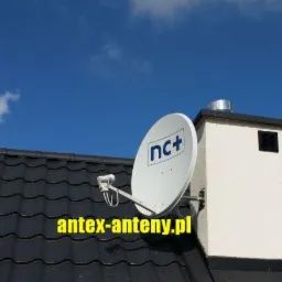 Biała antena satelitarna nc+ zamontowana na kominie domu z czarną dachówką na tle błękitnego nieba.