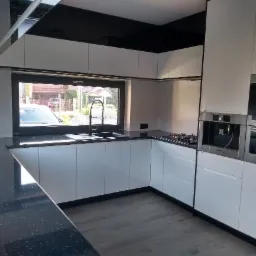 Nowoczesna kuchnia z białymi szafkami, czarnym blatem w kropki i czarnym sufitem podwieszanym, z widokiem na zewnątrz przez okno nad zlewem.