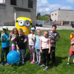 Grupa dzieci pozuje do zdjęcia z animatorem przebranym za Minionka na tle budynku podczas imprezy plenerowej.