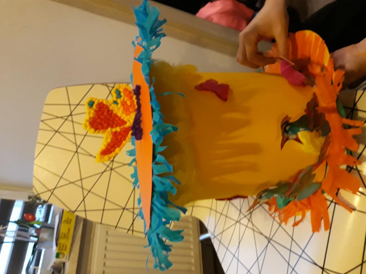 Ręcznie robiona, żółta piñata w trakcie dekorowania papierowymi elementami w kolorach pomarańczy, turkusu i czerwieni, widoczne dłonie osoby pracującej nad nią.