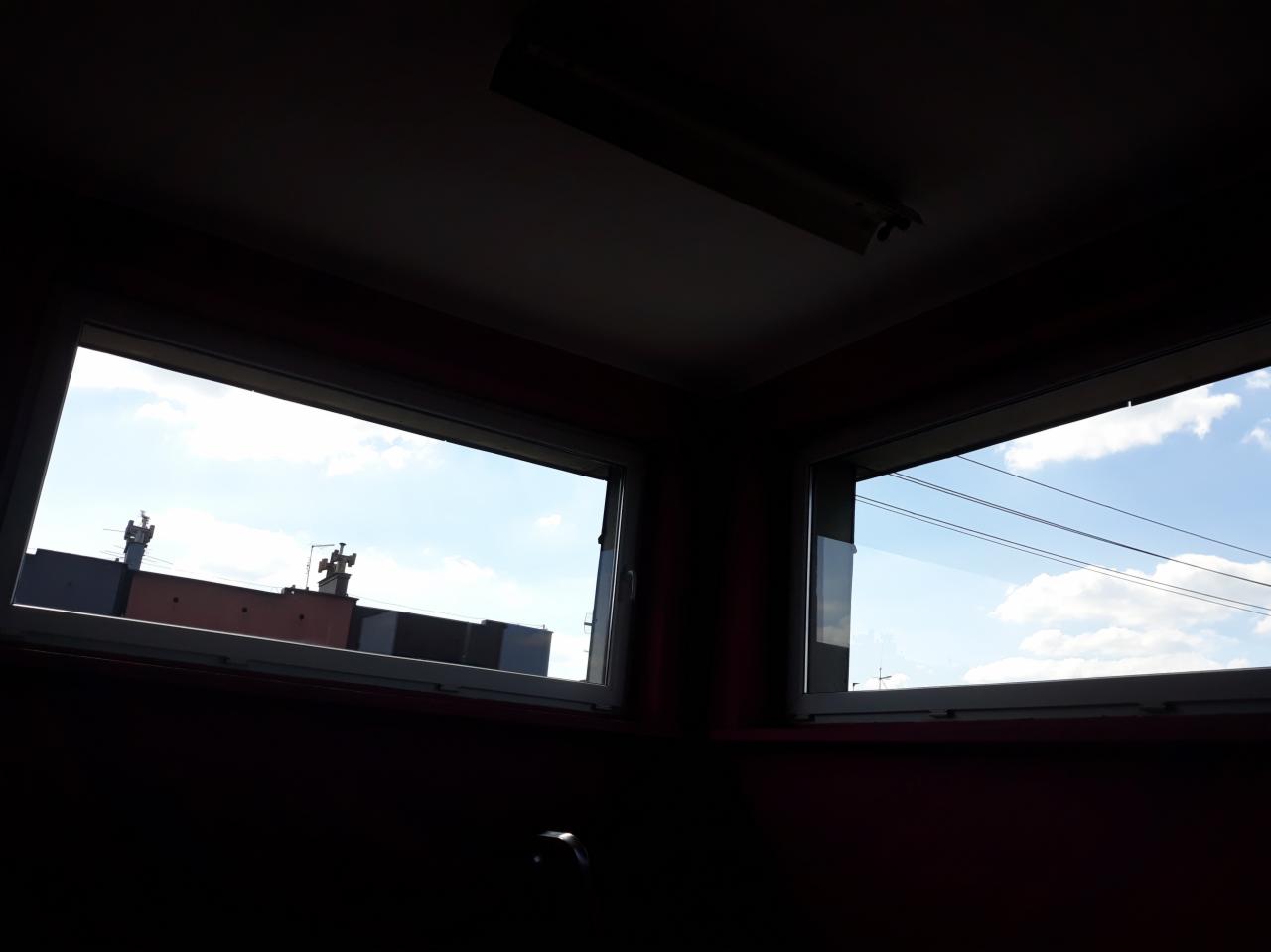 Dwa okna z widokiem na błękitne niebo i budynki z antenami, sfotografowane z ciemnego wnętrza.