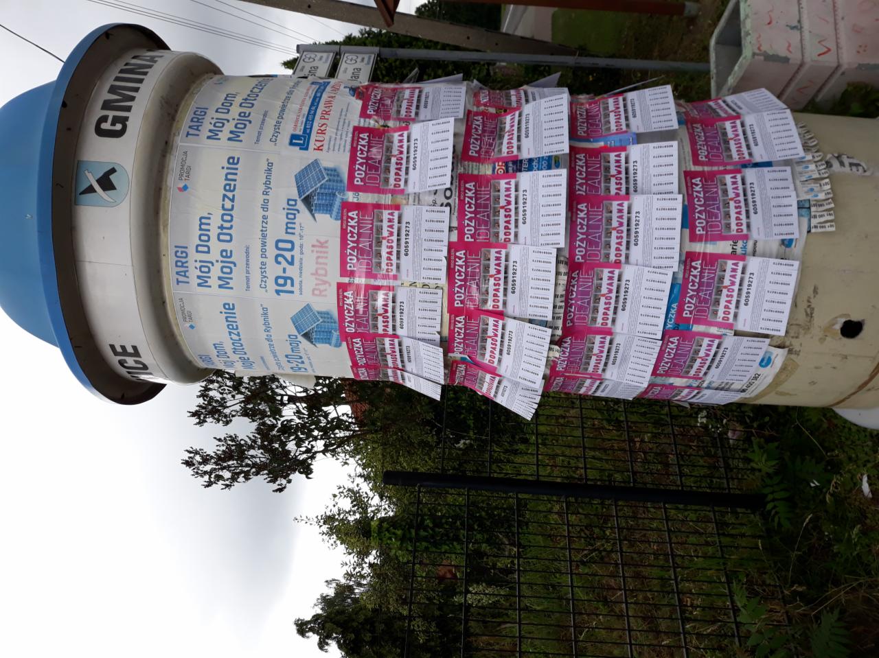 Ogłoszeniowy słup z naklejonymi plakatami reklamującymi pożyczki i targi lokalne, otoczony zielenią i ogrodzeniem.