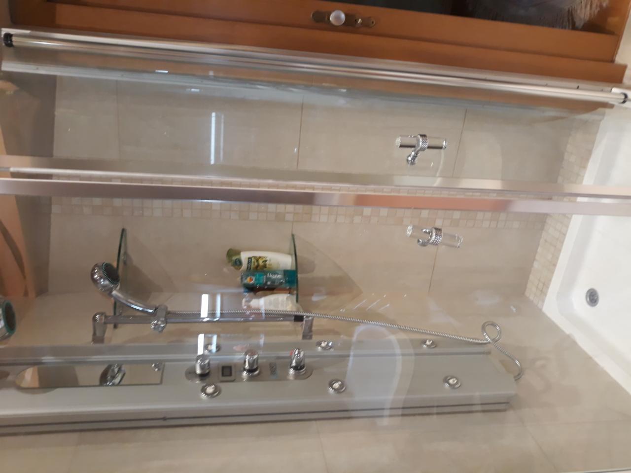 Szklana kabina prysznicowa z hydromasażem, widoczne dysze, słuchawka i półka z kosmetykami, jasne płytki ceramiczne z mozaiką.