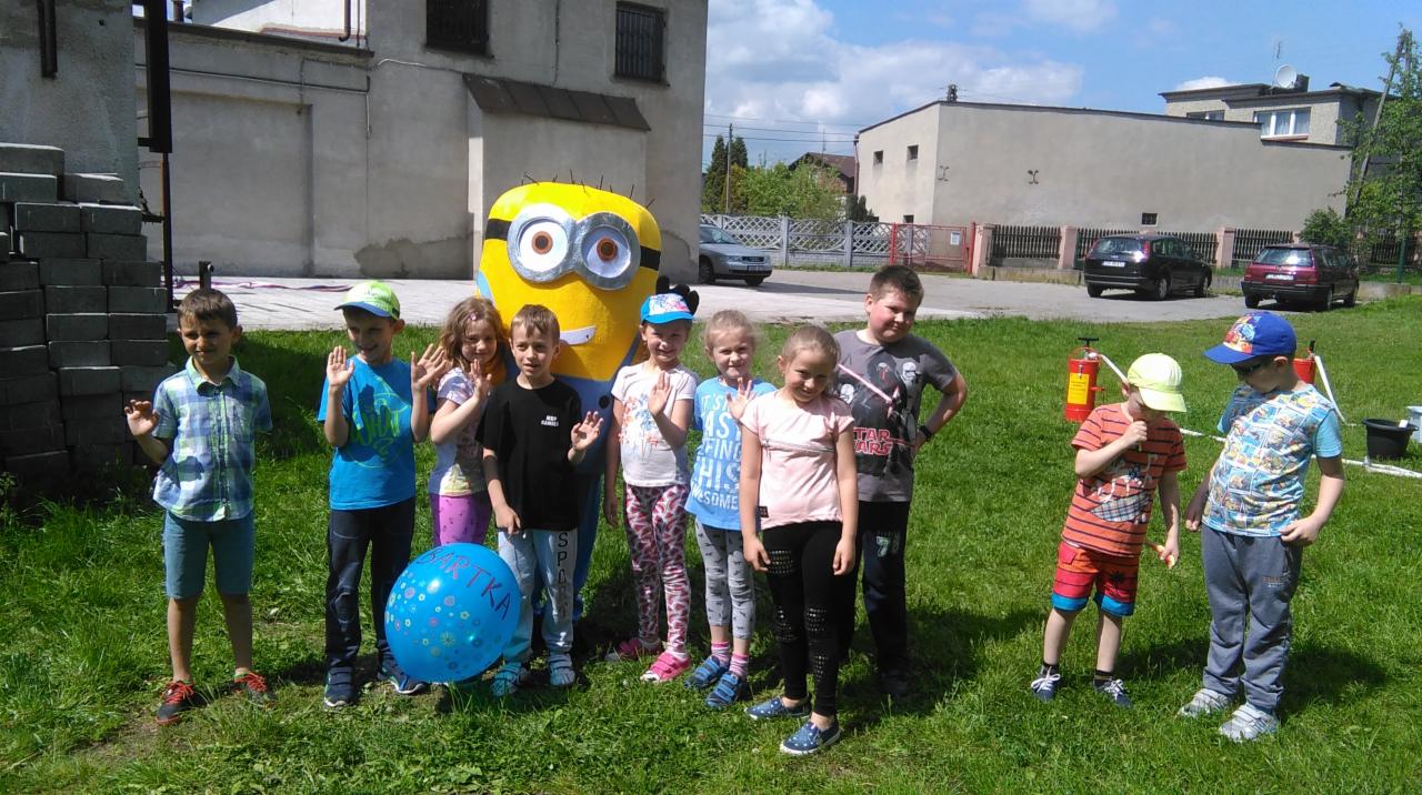 Grupa dzieci pozuje do zdjęcia z animatorem przebranym za Minionka na tle budynku podczas imprezy plenerowej.