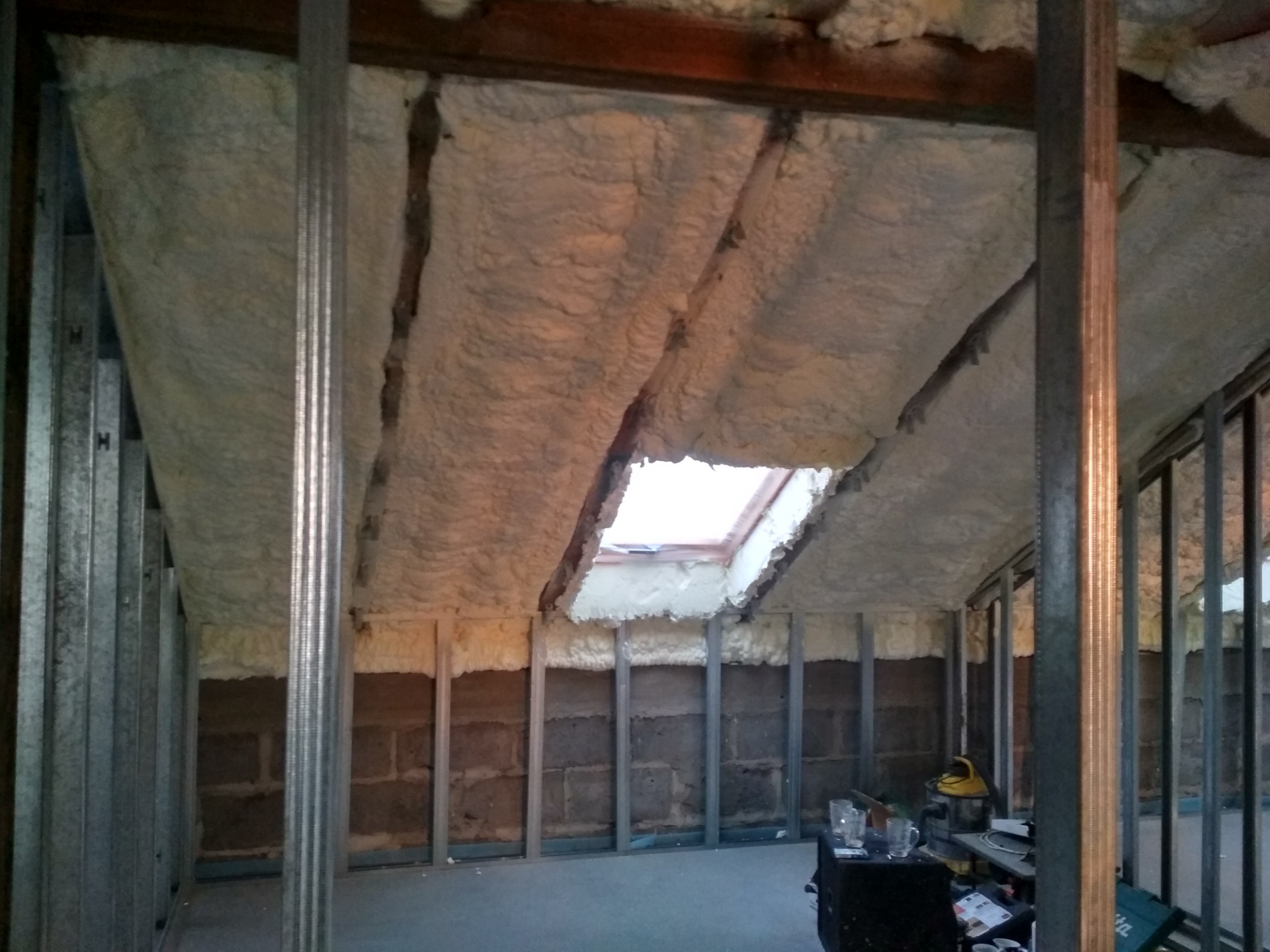 Poddasze z widoczną aplikacją izolacji na bazie pianki, z metalowymi profilami konstrukcyjnymi i oknem dachowym.