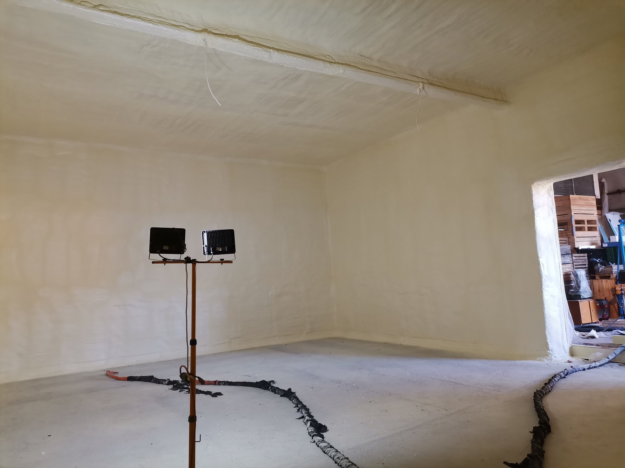 Wnętrze pomieszczenia po natrysku izolacji, widoczne lampy oświetleniowe na statywie i wąż do natrysku leżący na podłodze.