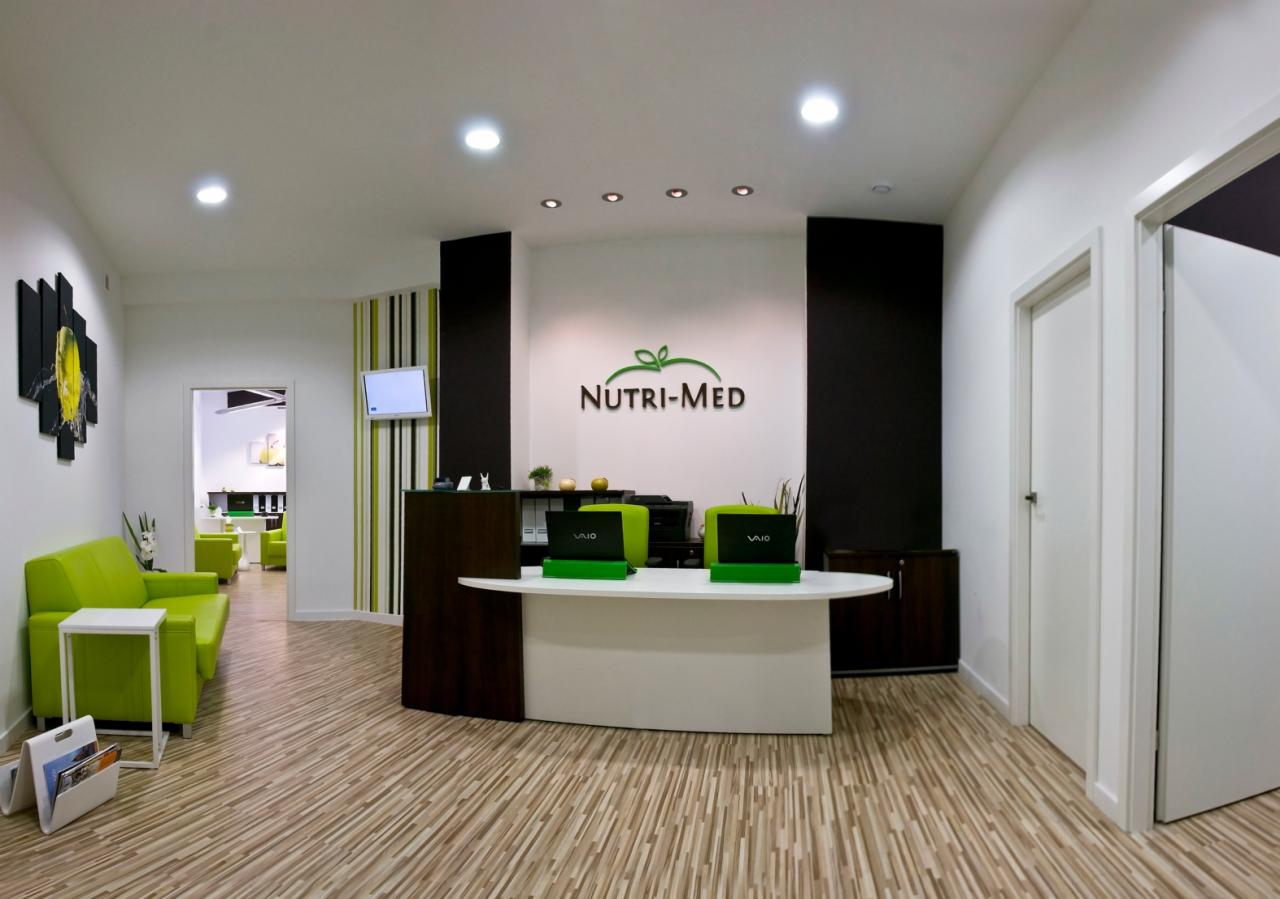 Recepcja gabinetu dietetycznego Nutri-Med z logo na ścianie, zielonymi akcentami i laptopami VAIO, widok na poczekalnię z zielonymi sofami.
