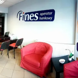 Wnętrze biura z logo 'Fines operator bankowy' na ścianie, czerwonymi fotelami, biurkiem z laptopem i regałem, oraz banerem reklamowym z hasłem 'Kredyty i pożyczki w jednym miejscu'.
