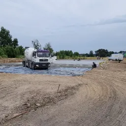 Przygotowanie terenu pod wylewkę: rozłożona folia, pracownicy, betoniarka i wyrównywanie powierzchni na placu budowy.