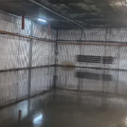 Świeżo wylana, mokra posadzka przemysłowa w hali z blachy falistej, widoczne odbicia ścian i instalacji w lustrzanej powierzchni, z fragmentem pomieszczenia socjalnego w tle.