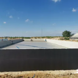 Świeżo wylana, jasnoszara płyta betonowa w dużej, otwartej przestrzeni, wyrównywana przez dwóch pracowników w oddali, z widoczną pompą do betonu po lewej stronie i czarną izolacją pionową...