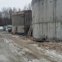 Budowa dwóch okrągłych, betonowych zbiorników z widocznym szalunkiem i drabiną, teren budowy z błotem i śniegiem, pochmurne niebo w tle.