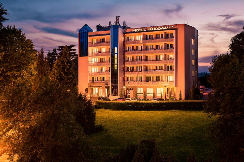Oświetlony nocą budynek Hotelu Kudowa z czterema gwiazdkami, widok z trawnika otoczonego drzewami.