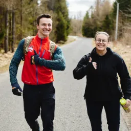 Uśmiechnięta para daje kciuki w górę podczas biegu po leśnej drodze; mężczyzna w czerwonej kurtce i z plecakiem, kobieta w czarnej bluzie trzyma bidon.