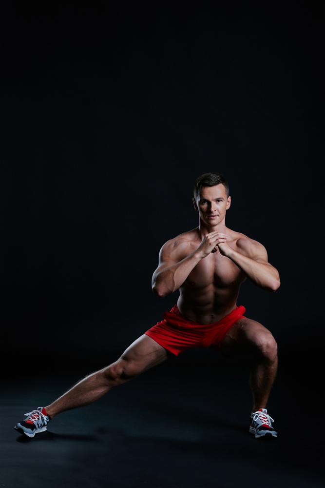 Mężczyzna w czerwonych spodenkach wykonuje wypad do boku na czarnym tle, prezentując umięśnioną sylwetkę.