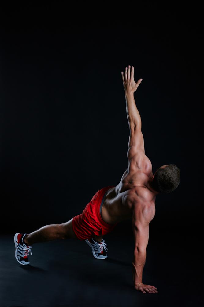 Mężczyzna w czerwonych spodenkach wykonuje ćwiczenie w podporze bokiem, unosząc jedną rękę w górę na tle czarnego tła.