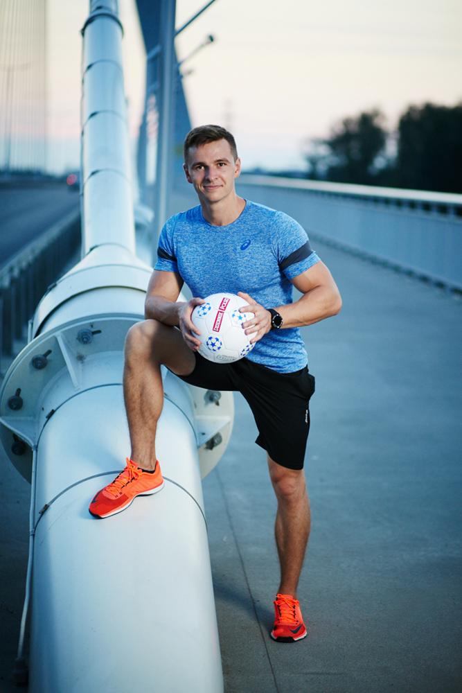 Uśmiechnięty mężczyzna w sportowym stroju pozuje z piłką na tle metalowej konstrukcji mostu.