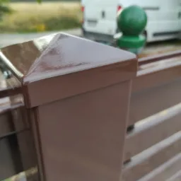 Fragment brązowego ogrodzenia panelowego z widocznym słupkiem zakończonym piramidką, w tle samochód dostawczy i zieleń.