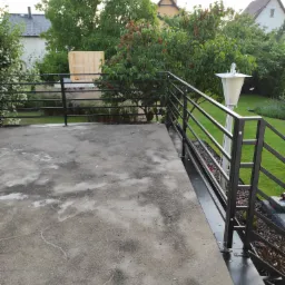 Czarna, metalowa balustrada z poziomymi szczeblami na betonowym tarasie, widok na ogród z drzewami i lampą ogrodową.