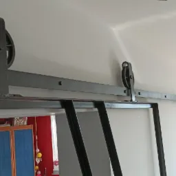 Metalowa konstrukcja przesuwnych drzwi w stylu loft z widocznymi rolkami i szyną mocowaną do sufitu, wnętrze pokoju z wzorzystą tapetą w tle.