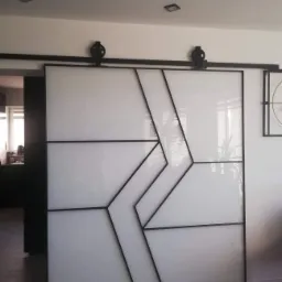 Nowoczesne, przesuwne drzwi wewnętrzne z białego szkła i czarnych metalowych profili, tworzące geometryczny wzór strzałki, zawieszone na czarnej szynie sufitowej z widocznymi rolkami.