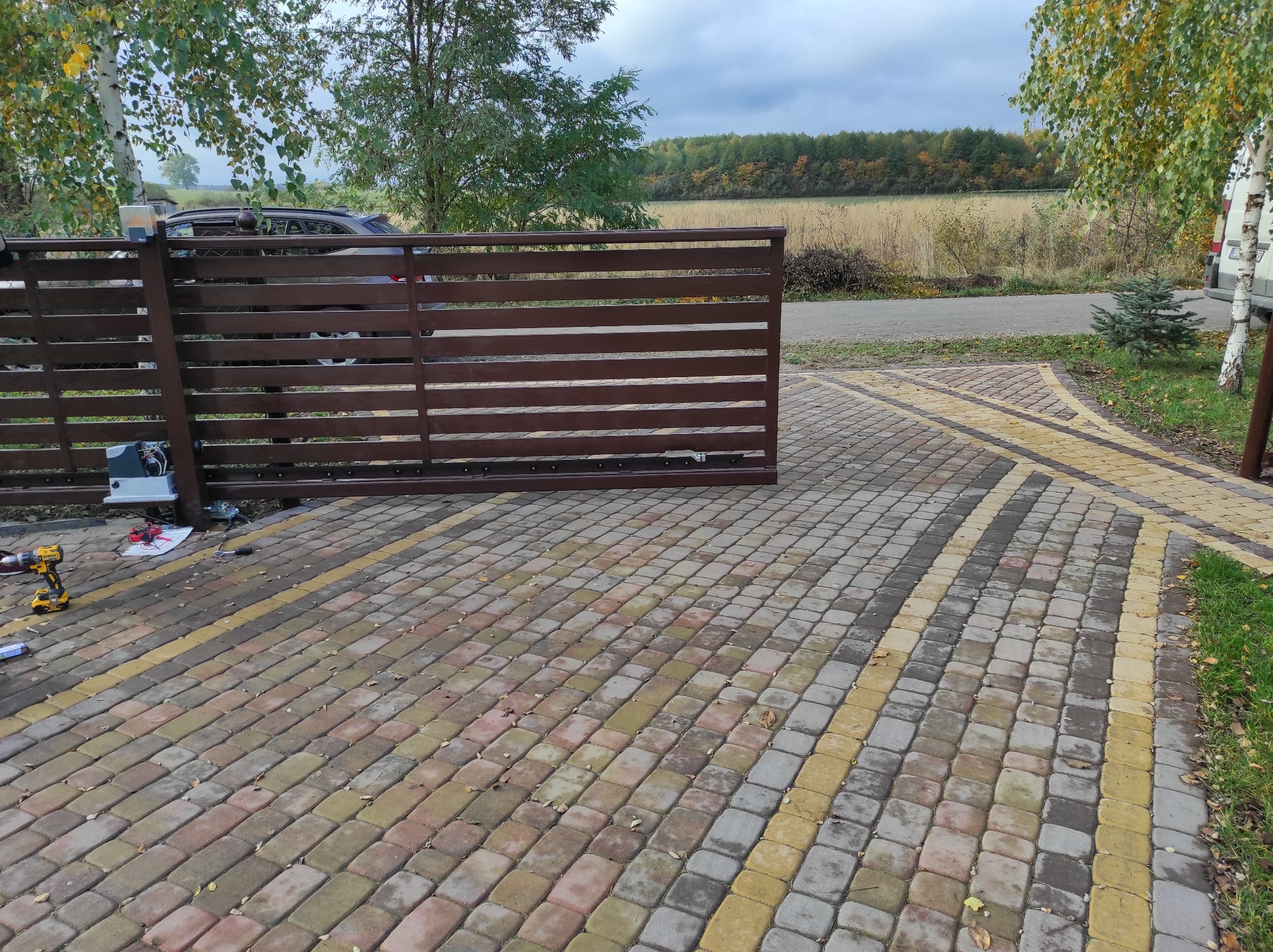 Brązowa brama wjazdowa z automatyką, częściowo otwarta, widoczna kostka brukowa, narzędzia montażowe i krajobraz polny w tle.