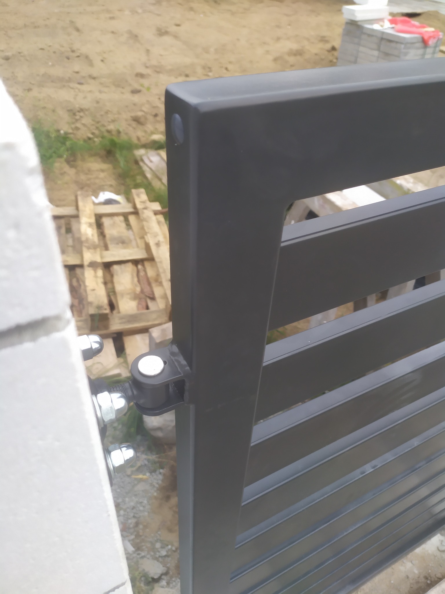 Fragment czarnej, poziomej bramy wjazdowej z metalowych profili, zamocowanej do białego słupka z betonu komórkowego za pomocą zawiasów i śrub.