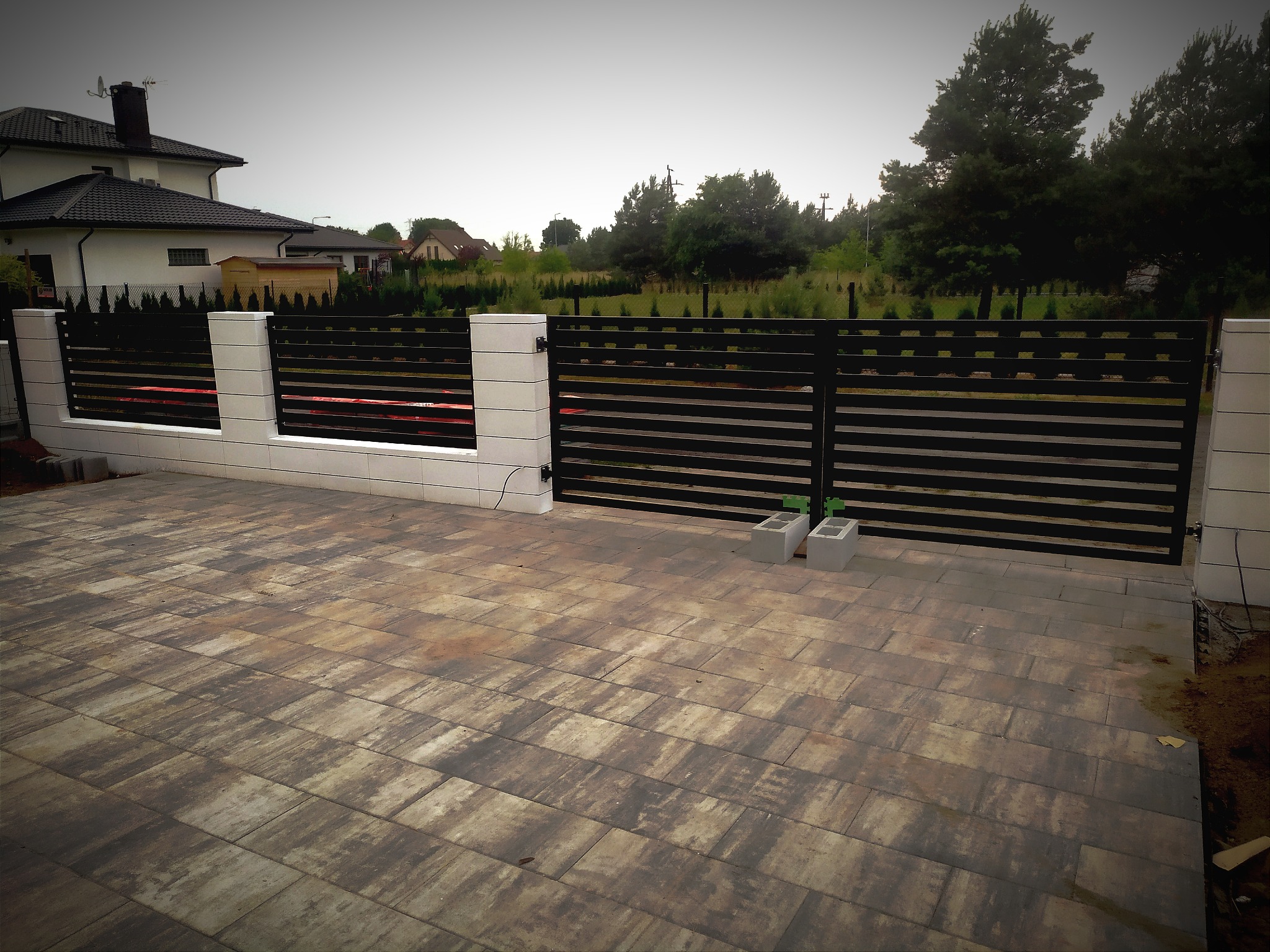 Nowoczesna, pozioma, czarna brama wjazdowa z białymi słupkami, widok z zewnątrz na brązową kostkę brukową i dom w tle.