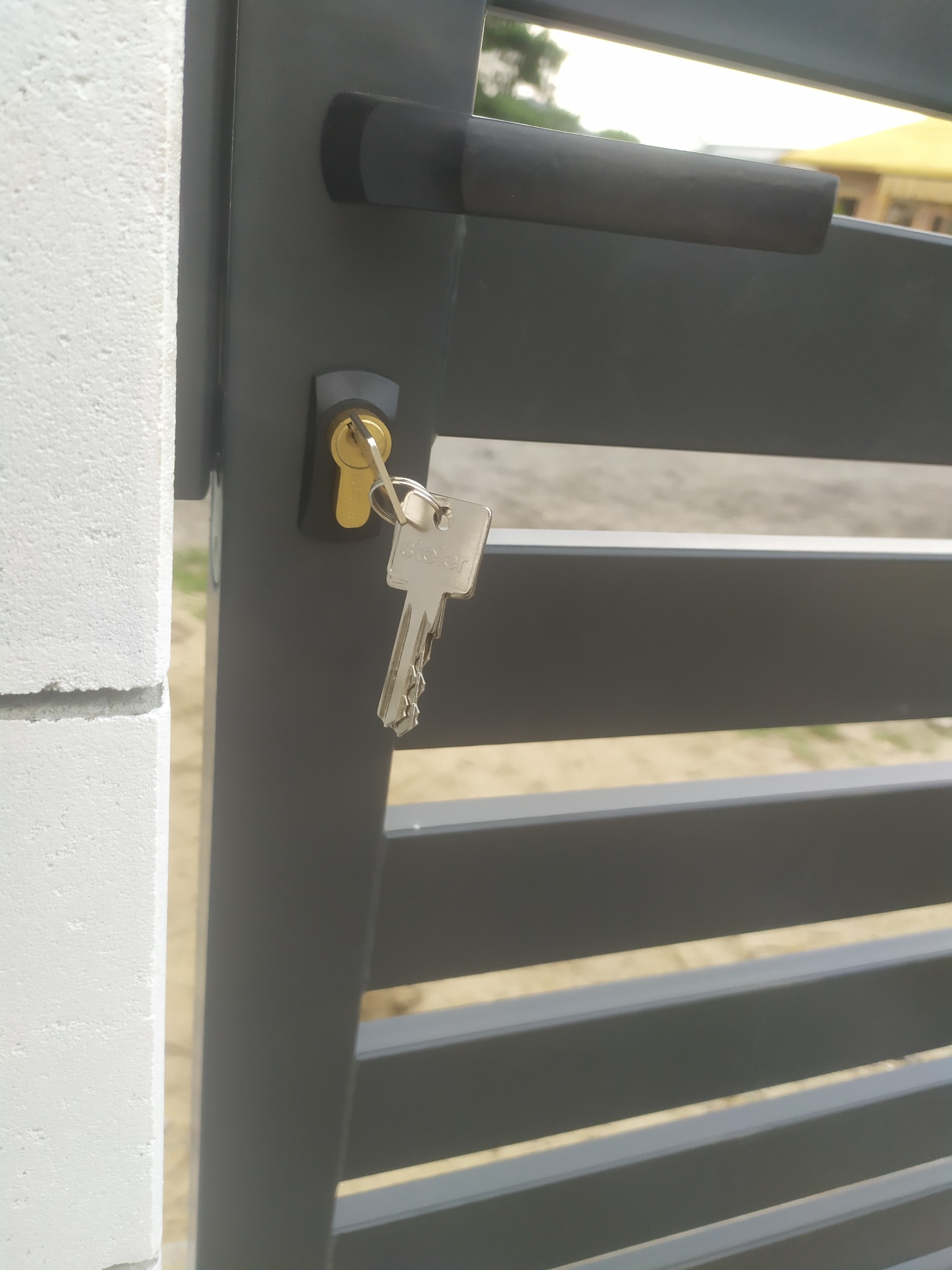 Szara furtka z poziomymi panelami, klucz w zamku, biały słupek ogrodzeniowy z betonu komórkowego po lewej stronie.