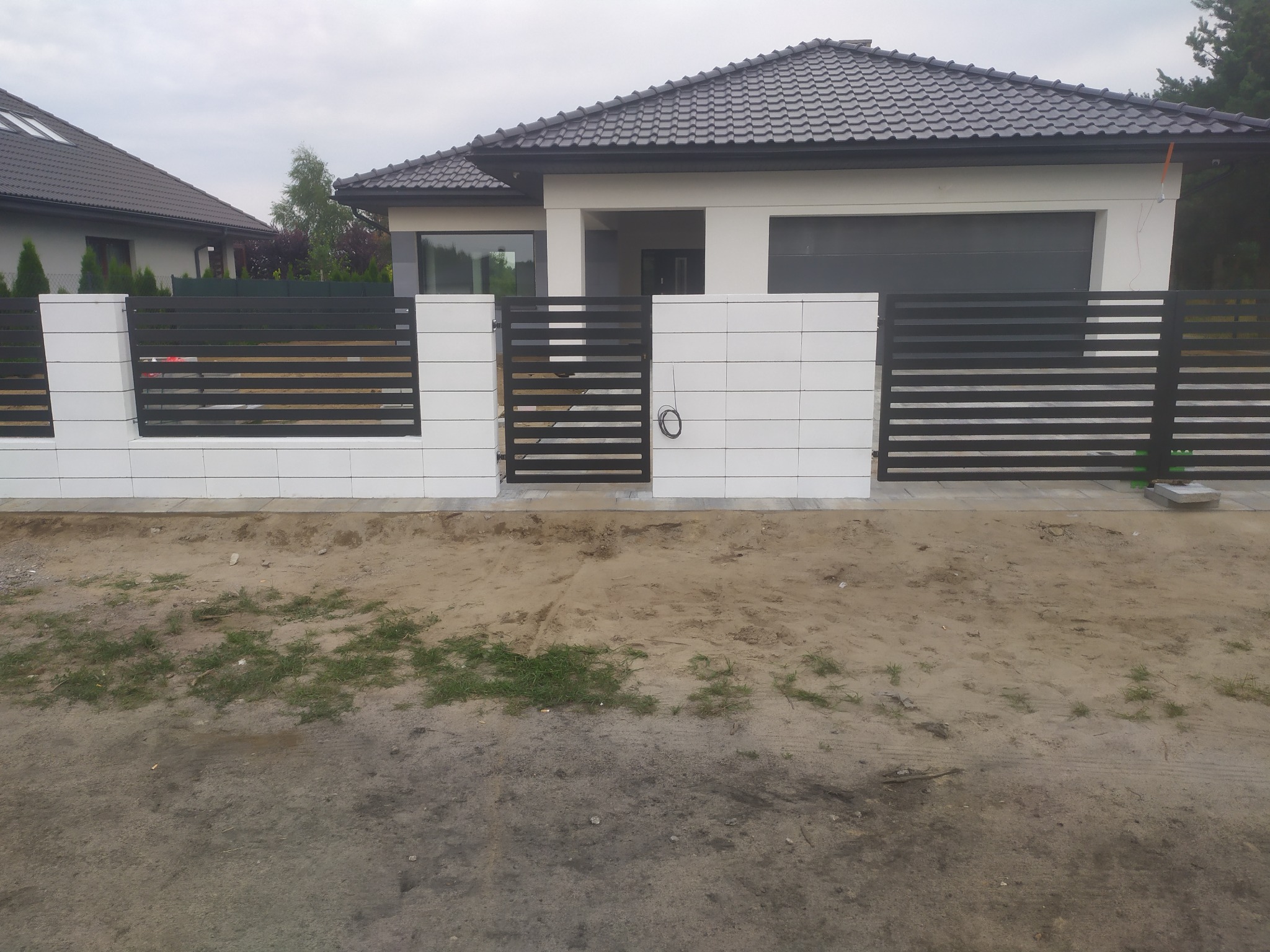 Nowoczesne ogrodzenie posesji z poziomych, grafitowych przęseł metalowych i białych, kwadratowych słupków z płytek elewacyjnych, na tle domu z szarą dachówką.