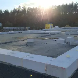 Przygotowany fundament pod budowę domu z bloczków Ytong, widoczne zabezpieczenie przeciwwilgociowe, w tle las i budynek mieszkalny w oddali, słoneczny dzień.