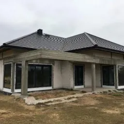 Dom w stanie surowym otwartym z czarną dachówką, widoczne betonowe elementy konstrukcyjne i duże okna, otoczony trawiastym terenem w pochmurny dzień.