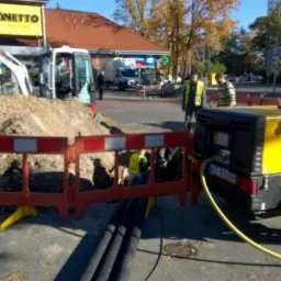 Żółty kompresor CompAir na przyczepie, rów z czarnymi rurami, bariera ochronna z czerwono-białymi pasami, koparka przed budynkiem sklepu, dwóch pracowników w tle.