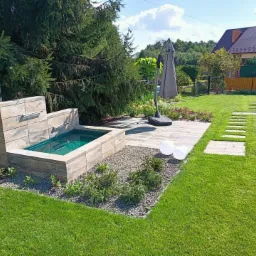 Ogród z małym oczkiem wodnym obudowanym kamieniem, trawnikiem, ścieżką z płyt i parasolem ogrodowym w tle domu.