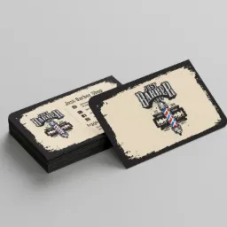 Stos wizytówek fryzjerskich z logo 'Just Barber Shop' z motywem tradycyjnej kolumny fryzjerskiej i ostrza brzytwy na beżowym tle z czarną obwódką, ułożonych na szarym tle.