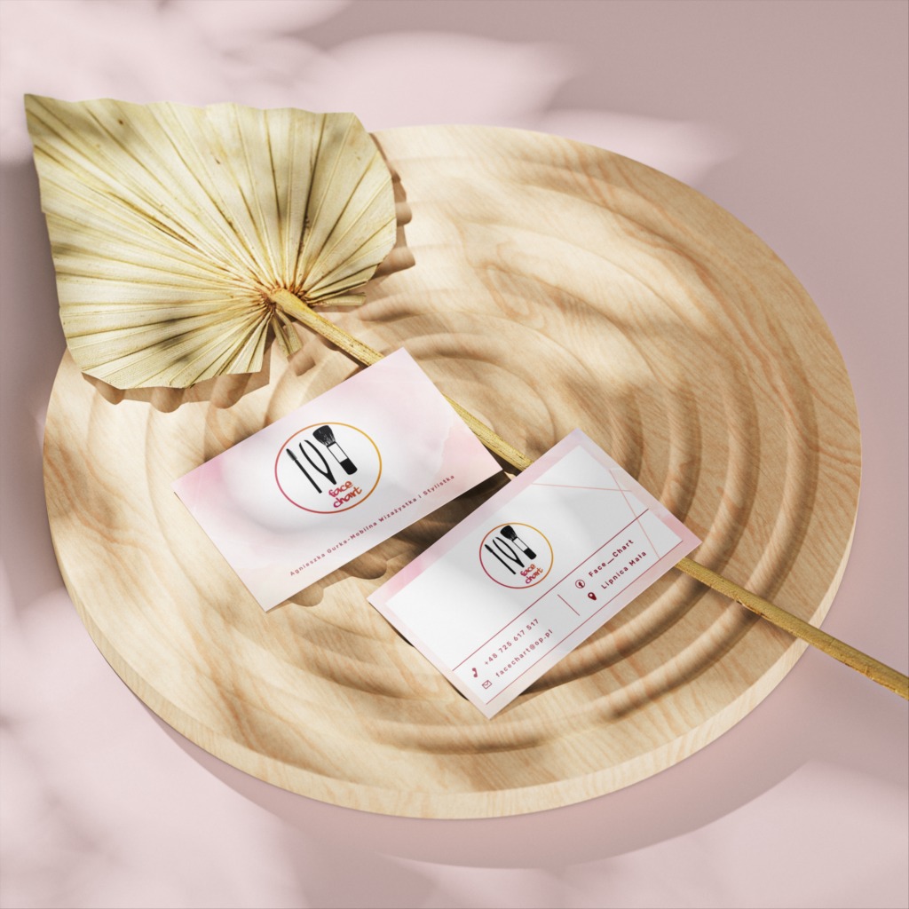 Dwie wizytówki salonu makijażu 'Face Chart' leżą na drewnianej tacy z suszonym liściem palmy w tle.