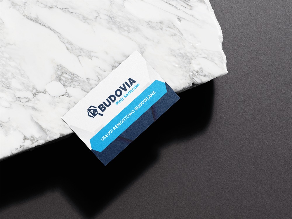 Wizytówka firmy Budovia Piotra Kazieczko oferującej usługi remontowo-budowlane, leżąca na marmurowej płycie na ciemnoszarym tle.