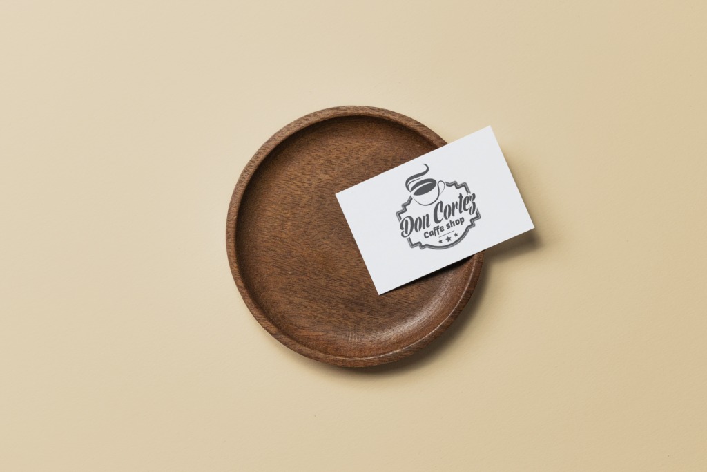 Wizytówka kawiarni Don Cortez leży na ciemnobrązowym drewnianym talerzyku na beżowym tle. Logo kawiarni zawiera filiżankę kawy i nazwę firmy.