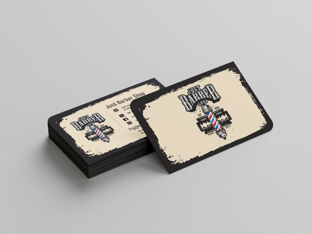 Stos wizytówek fryzjerskich z logo 'Just Barber Shop' z motywem tradycyjnej kolumny fryzjerskiej i ostrza brzytwy na beżowym tle z czarną obwódką, ułożonych na szarym tle.