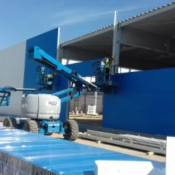 Montaż niebieskich paneli elewacyjnych na konstrukcji stalowej hali przemysłowej z użyciem podnośników koszowych, widoczne zapakowane płyty warstwowe