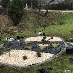 Weles Patryk Macięga - Aranżacja ogrodu z białym i czarnym grysem, obrzeża z kostki, niskie krzewy ozdobne. Na trawie widoczne narzędzia ogrodnicze i resztki materiałów.